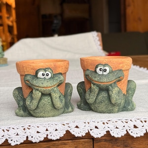 May include: Two terracotta-coloured plant pots, each featuring a textured green frog figure attached to the front. The frogs have wide, smiling mouths, large white eyes with black pupils, and hands resting under their chins against the pot.