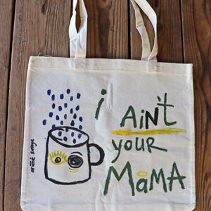 Könnte beinhalten: Eine weiße Canvas-Tragetasche mit handgemaltem Design. Das Design zeigt eine blaue Kaffeetasse mit einem gelben Auge und einem grün-gelben Heiligenschein. Der Text "I Ain't Your Mama" ist in schwarzer, grüner und gelber Farbe geschrieben.