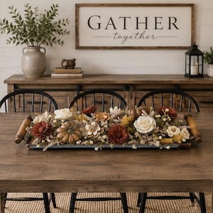 May include: A rectangular wooden dining table with a trestle base and four black spindle-back chairs. Centered on the table is a long metal tray with wooden handles, filled with an arrangement of artificial flowers, foliage, and small pumpkins.
