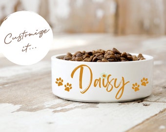May include: White ceramic pet bowl filled with dry dog food. The bowl is personalized with the name "Daisy" in gold script, paw prints, and a small floral accent. The text "Customize it..." is in a white circle.