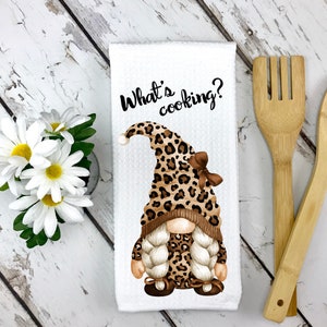 May include: White kitchen towel with a gnome wearing a leopard print hat and bow. The text "What's cooking?" is printed above the gnome. Wooden spoon and spatula are next to the towel. Daisies are in a small vase.