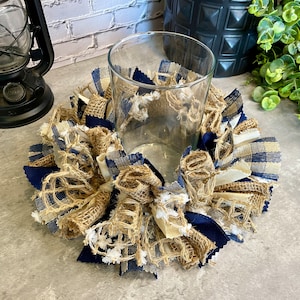 May include: A clear glass candle holder surrounded by a decorative burlap and fabric wreath. The wreath features navy blue, beige, and white fabric accents. The candle holder is on a gray surface.