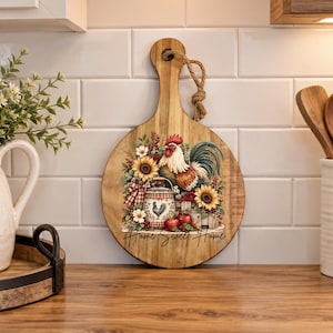 May include: A round wooden cutting board with a handle and a jute rope loop. The board features a printed illustration of a rooster, sunflowers, red apples, and a small bucket, with the text Home Sweet Home written in black script.