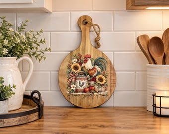 May include: A round wooden cutting board with a handle and a jute rope loop. The board features a printed illustration of a rooster, sunflowers, red apples, and a small bucket, with the text Home Sweet Home written in black script.