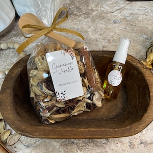May include: A wooden bowl holds a clear bag of potpourri with a cinnamon stick and a tag that reads "Cinnamon and Vanilla." A small spray bottle and a jar of dried botanicals are also present. The items are arranged on a surface.
