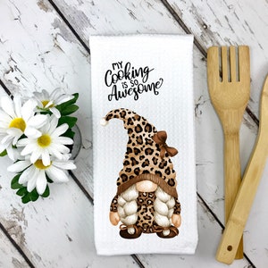 May include: White kitchen towel with a gnome wearing a leopard print hat and outfit. The text "MY COOKING IS SO AWESOME" is printed above the gnome. Wooden cooking utensils and white daisies are also in the image.
