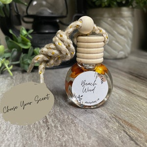 May include: A small, clear glass bottle with a wooden cap and a knotted rope. The bottle contains a liquid with orange and yellow accents and a label that reads "Beach Wood." A tag with "Choose Your Scent" hangs from the rope.