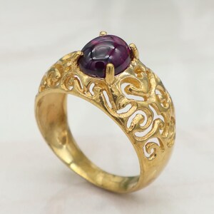 Può includere: Un anello color oro con un design traforato ornato. L'anello presenta una grande gemma ovale, rosso scuro. L'anello è posto su una superficie bianca con uno sfondo verde sfocato.