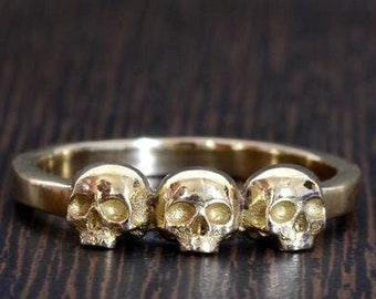 May include: A gold ring with three skull designs. The ring is on a dark wood surface.