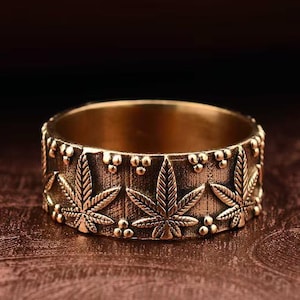 Könnte beinhalten: Ein goldfarbener Ring mit einem sich wiederholenden Muster aus Cannabispflanzenblättern und kleinen perlenartigen Akzenten. Der Ring besteht aus glänzendem Metall und hat eine glatte Innenseite.
