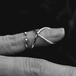 May include: A silver ring with an X-shaped design on the finger. A smaller ring with two silver spheres is also visible. The jewellery is set against a dark background.
