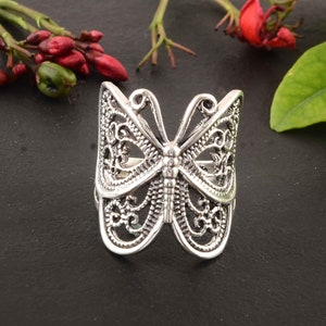 Puede incluir: Un anillo de plata con forma de mariposa con intrincados detalles de filigrana. El anillo es ancho y tiene un diseño delicado y calado.