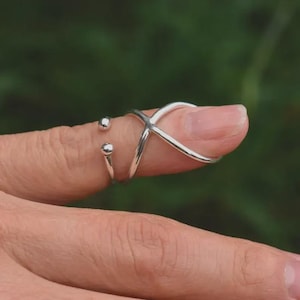 Könnte beinhalten: Ein silberfarbener Fingerring mit einem X-förmigen Design und zwei kleinen, runden Akzenten. Der Ring wird an einem Finger getragen, mit einem verschwommenen grünen Hintergrund.