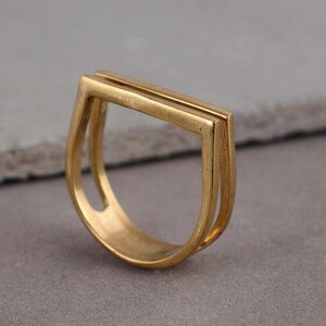 May include: A gold-toned ring with a double-layered, rectangular design. The ring is open on one side and has a rounded shape.