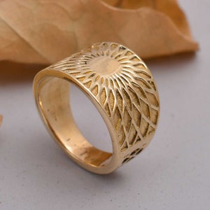May include: A gold ring with a wide band featuring a geometric design that resembles a sunburst.