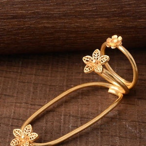 Könnte beinhalten: Ein goldfarbenes Haaraccessoire mit floralem Design. Das Accessoire hat eine gebogene, längliche Form mit drei dekorativen Blumenakzenten. Die Blumen haben detaillierte, durchbrochene Blütenblätter. Das Stück ist vor einem dunklen Holzgrund platziert.
