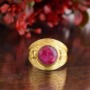 May include: A gold-colored ring with a large, oval, ruby-red gemstone. The ring has intricate detailing around the band and the gemstone setting. The background is blurred, with hints of red and brown.