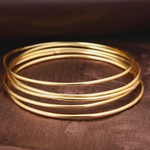 May include: A stack of five gold bangles. The bracelets are thin and round, with a smooth, shiny surface. They are stacked together on a brown surface, creating a layered effect. The bangles are a warm, golden color.