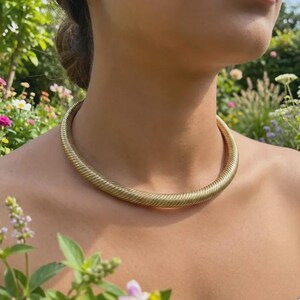 Puede incluir: Un collar dorado y acanalado. El collar es grueso y se ajusta al cuello. El fondo presenta un jardín con flores y vegetación.