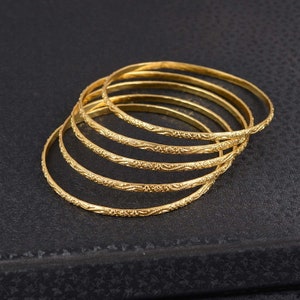 Op de afbeelding: Een stapel van zes goudkleurige armbanden met een gegraveerd bloemmotief. De armbanden liggen op een donkere ondergrond, met het licht dat weerkaatst op het gepolijste metaal. De armbanden zijn een klassiek sieraad.