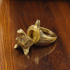 May include: A detailed brass ring with a wolf head design. The ring features a textured finish and a setting for a gemstone. The wolf's head is intricately carved, showcasing the animal's features. The ring is displayed on a wooden surface.