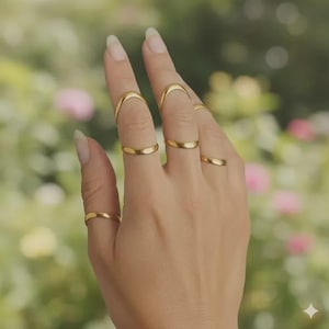 May include: A hand wearing several gold rings. The rings are different styles, including simple bands and curved designs. The background is a soft, blurred view of greenery and flowers.