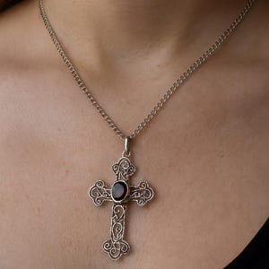 Puede incluir: Collar con colgante de cruz de filigrana plateada con una piedra ovalada oscura en el centro. La cruz presenta adornos de volutas y cuelga de una delicada cadena. El collar se muestra sobre un fondo oscuro.