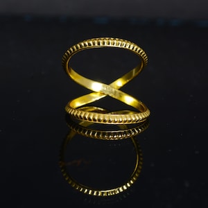 May include: A gold ring with an overlapping design. The ring features a textured band with small, vertical lines. The ring's design forms an X shape. The ring is displayed on a reflective black surface.