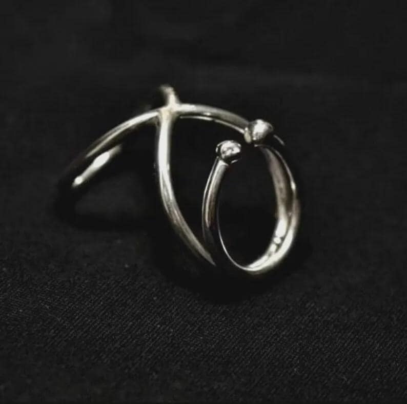 May include: A silver ring with a distinctive design. The ring has a curved band that encircles the finger, with two small, round embellishments. The ring is set against a black background.