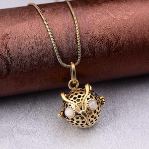 Può includere: Una collana con pendente a forma di gufo dorato. Il pendente a forma di gufo presenta un design traforato dettagliato con due accenti bianchi e rotondi per gli occhi. La catena della collana è una sottile catena a serpente dorata. Lo sfondo è una superficie marrone strutturata.