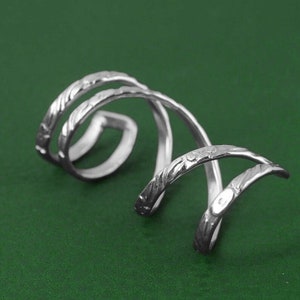 Könnte beinhalten: Silberner Ohrring mit einem gedrehten, strukturierten Design. Der Ohrring besteht aus drei miteinander verbundenen Metallbändern.