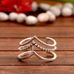 Könnte beinhalten: Ein silberfarbener Ring mit einem einzigartigen Design. Der Ring hat drei Bänder, wobei das obere und mittlere Band mit kleinen, runden Perlen verziert sind. Das untere Band hat eine V-Form. Der Ring liegt auf einer Holzoberfläche.