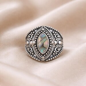 May include: A silver-toned ring with an oval, iridescent abalone shell center stone. The ring features intricate, detailed filigree and beadwork around the stone and band, creating a vintage or bohemian aesthetic.
