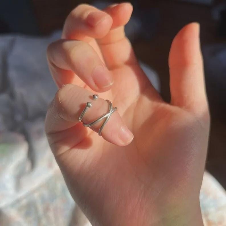 May include: A silver ring with a distinctive design is worn on a finger. The ring has two small spheres and a crisscross pattern. The ring is made of a shiny metal and is a piece of jewellery.