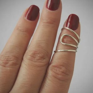 May include: A silver-toned finger ring with a unique, flowing design. The ring wraps around the finger with a series of curved lines, creating an abstract and modern look. The image shows the ring on a finger with dark red nail polish.