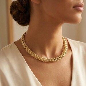 Puede incluir: Un collar tejido de color dorado. El collar es de estilo gargantilla, con un diseño grueso y texturizado. La joya se lleva alrededor del cuello, y la imagen es un primer plano del collar y parte de la persona que lo lleva.