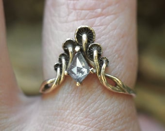 May include: A gold ring with a clear, faceted gemstone set in the center. The ring features a design of stylized mushrooms surrounding the stone.