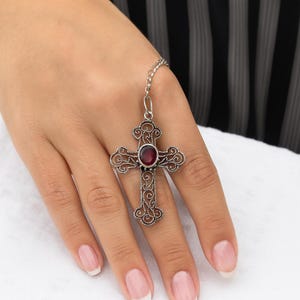 Op de afbeelding: Zilverkleurige kruisring met een sierlijk filigraanontwerp en een ovale, dieprode edelsteen in het midden. De ring is verbonden met een fijne ketting. De achtergrond is een zacht witte ondergrond en een zwart-wit gestreepte stof.