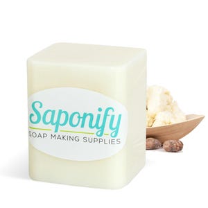 May include: A white block of shea butter with a label that reads "Saponify Soap Making Supplies". There are shea nuts and a wooden bowl with shea butter in the background.