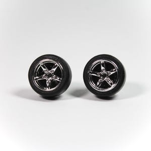 May include: Two miniature car wheels with black tires and shiny, silver five-spoke rims. The wheels are set against a plain white background, highlighting their design and detail. These wheels are likely for a model car or a similar small-scale project.