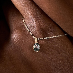 Puede incluir: Primer plano de una persona que lleva un collar y pendientes de oro. El collar presenta un pequeño colgante ovalado con una piedra preciosa verde oscuro. Los pendientes a juego también son de color verde oscuro.