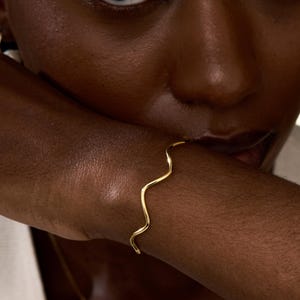 Pode incluir: Uma pulseira dourada com um design ondulado. A pulseira é usada no pulso de uma pessoa. A imagem é um close-up, destacando o design da joia e o tom de pele da pessoa. O design da pulseira é simples e elegante.