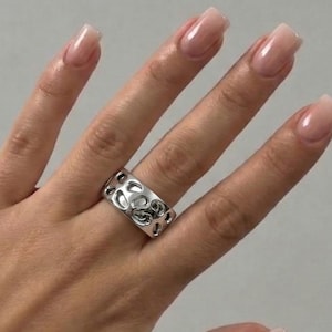 Pode incluir: Um anel prateado com um design floral é usado num dedo. O anel tem detalhes vazados, e a mão é mostrada contra um fundo neutro. A pessoa está usando uma jaqueta de couro marrom.