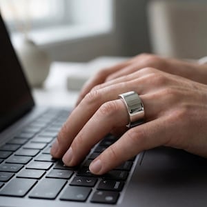 May include: A silver ring with a flat top and a wide band is worn on a finger as a person types on a laptop keyboard. The ring has a polished, reflective surface. The background is blurred, suggesting an office or home setting.
