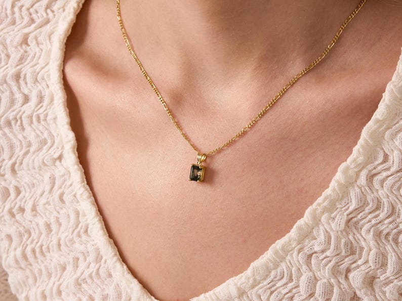 May include: A gold chain necklace with a small, square-shaped pendant featuring a dark green gemstone.