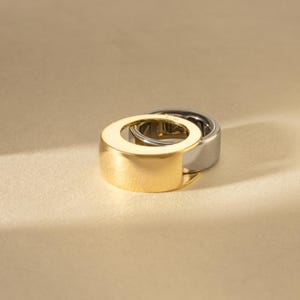 May include: Two modern rings, one gold and one silver, are stacked together. The gold ring is in front, partially obscuring the silver ring behind it. The rings have a minimalist design with a flat, wide band and a circular opening in the center.