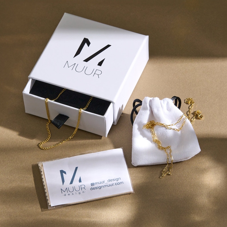 K&ouml;nnte beinhalten: Eine goldene Kettenkette in einer wei&szlig;en Box mit einer schwarzen Schublade. Die Box tr&auml;gt das Logo "M/A MUUR design" auf der Oberseite. Die Kette befindet sich auch in einem wei&szlig;en Beutel mit Kordelzug. Ein wei&szlig;es Reinigungstuch mit dem Logo "M/A MUUR design @muur_design designmuur.com" liegt auf dem Tisch.