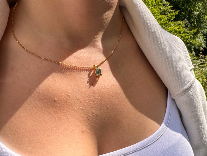 May include: A gold chain necklace with a small, square-shaped pendant featuring a dark green gemstone.