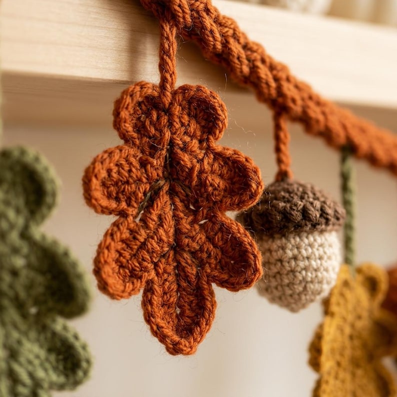 May include: A crochet garland featuring hanging autumn-themed elements including a rust-colored oak leaf, a beige acorn with a dark brown cap, and additional leaves in olive green and mustard yellow, all attached to a thick, braided yarn cord.