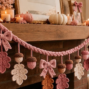 May include: A decorative garland featuring crocheted elements in shades of pink, cream, and brown. The garland consists of a braided pink cord adorned with hanging crocheted bows, oak leaves, and acorns, creating a textured, seasonal display.